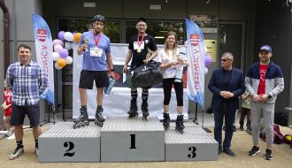 Trzy dorosłe osoby na rolkach i z medalami stoją na podium przed RCK w Drzewicy. Zwycięzca trzyma czarną torbę, zdobywca drugiego miejsca jest w kasku, a kobieta z trzeciego miejsca trzyma zwiniętą matę. Po bokach asystują Wiceprezes LGD „Nad Drzewiczką”, Burmistrz Drzewicy i Prezes DCW „Ofiarna dłoń. W tle ścianka reklamowa i balony.