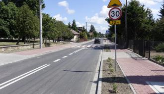 Nowy odcinek asfaltowej drogi z chodnikiem z czerwonej kostki. Na pierwszym planie pas zieleni oraz pionowe znaki drogowe: ograniczenie prędkości do 30 km/h i próg zwalniający.
