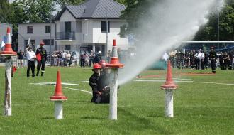 Ćwiczenie bojowe na trawiastym boisku podczas zawodów strażackich. Dwóch strażaków w hełmach i czarnych mundurach klęczy na trawie i kieruje silny strumień wody z węża w stronę ustawionych na białych słupkach czerwonych pachołków. W tle sędziowie z flagami, publiczność oraz piętrowy budynek mieszkalny.
