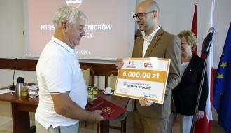 Mężczyzna w jasnobrązowym garniturze wręcza starszemu mężczyźnie w białej koszulce polo duży, symboliczny czek na kwotę 6000,00 zł na realizację projektu „WYPRAWA PO ZDROWIE”.