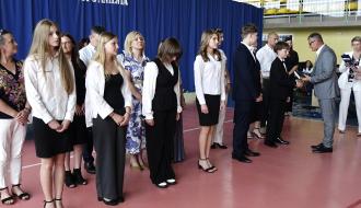 Grupa uczniów w strojach galowych stoi w rzędzie na sali gimnastycznej, podczas gdy Burmistrz Drzewicy w szarym garniturze wręcza dyplom i składa gratulacje jednemu z chłopców.