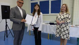  Burmistrz Drzewicy i Pani Dyrektor SP w Idzikowicach w czarno-białej sukience stoją z wyróżnioną uczennicą trzymającą otwarty dyplom.