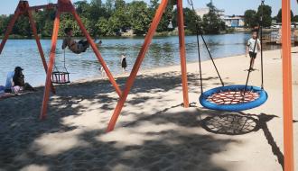 Nowoczesne huśtawki, w tym typu bocianie gniazdo, na piaszczystej plaży nad jeziorem. W tle widoczna woda i kąpiący się ludzie.