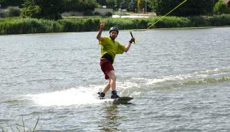 Mężczyzna w kasku, zielonej koszulce i bordowych spodenkach płynie na desce wakeboardowej, trzymając się orczyka. Lewą ręką wykonuje gest zwycięstwa (V), zostawiając za sobą białą smugę spienionej wody. W tle widoczny jest zielony, zarośnięty brzeg oraz dachy domów.