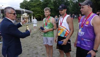 Ceremonia wręczenia nagród. Burmistrz Drzewicy w garniturze przekazuje puchar za pierwsze miejsce trzem zawodnikom z medalami. W tle drzewa.