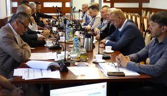 Uczestnicy sesji Rady Miejskiej w Drzewicy siedzą po obu stronach stołu konferencyjnego i biorą udział w obradach. Na stołach znajdują się dokumenty, mikrofony oraz napoje.