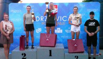 Dekoracja podium kobiet open: trzy biegaczki na stopniach 1, 2 i 3 z pucharami oraz torbami prezentowymi, w asyście organizatorów.