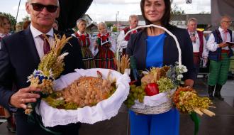 Burmistrz Drzewicy w garniturze oraz Przewodnicząca Rady Miejskiej w Drzewicy w niebieskim stroju prezentujący dożynkowy chleb oraz kosz z owocami i warzywami.