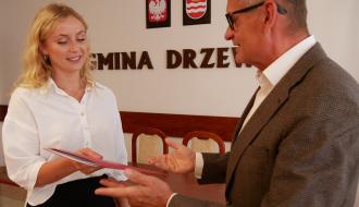 Burmistrz Drzewicy w garniturze i okularach gestykuluje dłońmi podczas przekazywania czerwonej teczki z aktem mianowania nauczycielce w białej koszuli.