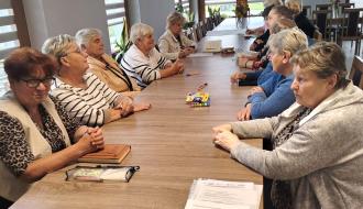 Grupa osób starszych siedząca naprzeciwko siebie przy długim stole w świetlicy w Werówce z kartkami i przyborami do pisania. 