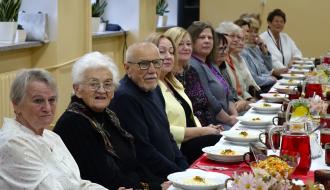 Seniorki i seniorzy siedzą przy długim, odświętnie nakrytym stole podczas poczęstunku. Na stole znajdują się talerze z jedzeniem, dzbanki z napojami oraz wazony z kwiatami. Uczestnicy spotkania patrzą w stronę obiektywu i uśmiechają się. 