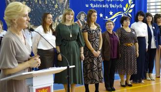 Pani Dyrektor SP w Radzicach Dużych przy mównicy z herbem Gminy Drzewica przemawia do mikrofonu. Z tyłu w szeregu stoją nauczycielki i nauczyciele. W tle znajduje się niebieska dekoracja rocznicowa. 