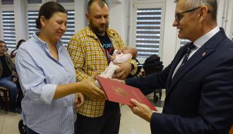 Burmistrz Drzewicy w garniturze wręcza pamiątkowy folder rodzicom trzymającym niemowlę na rękach. 