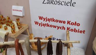 Potrawy regionalne – wędliny, miód i wypieki – od lokalnych producentów. Wystawka w ramach akcji „Kupuj świadomie”. W tle napis: „Zakościele Wyjątkowe Koło Wyjątkowych Kobiet