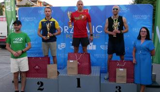 Na podium z pucharami i nagrodami trzej najlepsi biegacze w kategorii open mężczyzn. Po lewej przy podium stoi Zastępca Burmistrza Drzewicy Dominik Niemirski, po prawej Dyrektor Banki Spółdzielczego w Przysusze – Oddziału w Drzewicy Ewa Jakubczyk