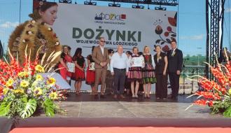 Na scenie Dożynek Województwa Łódzkiego trzyosobowa reprezentacja KGW Idzikowice tuż po odbiorze promesy z nagrodą za drugie miejsce. Obok po lewej Zastępca Burmistrza Drzewicy Dominik Niemirski, po prawej Marszałek Województwa Łódzkiego Joanna Skrzydlewska i Członek Zarządu Województwa Łódzkiego Artur Bagieński