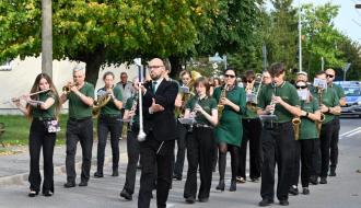  Orkiestra Miejska z Opoczna w zielonych koszulkach podczas marszu ulicznego. Na czele kapelmistrz w garniturze i okularach dyrygujący buławą, wokół niego muzycy z fletami, trąbkami, saksofonami i klarnetami.