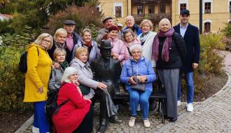 Grupa seniorów i uczestników wyjazdu pozuje do zdjęcia na tle historycznego budynku w parku, pozując wokół rzeźby siedzącej postaci Bolesława Prusa.