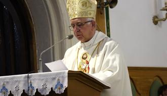 Arcybiskup Wacław Depo w mitrze i okularach stoi przy ambonie w kościele, czytając teksty liturgiczne z kartki papieru umieszczonej na mikrofonie.