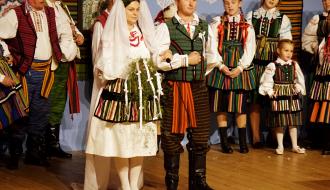 Para młoda w tradycyjnych strojach weselnych stoi w centralnej części sceny podczas widowiska obrzędowego Domaszna. Panna młoda ubrana jest w białą suknię z ozdobnym fartuchem i welonem, a pan młody w kamizelkę i wzorzyste spodnie. Obok stoją inni członkowie zespołu folklorystycznego i dzieci.