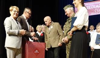 Trzyosobowa reprezentacja Dziennego Domu Senior+ w Drzewicy odbiera na scenie upominek rzeczowy z rąk Burmistrza Drzewicy Janusza Reszelewskiego. Obok Burmistrza stoi Jolanta Chełmińska - Pełnomocnik Wojewody Łódzkiego do spraw polityki senioralnej