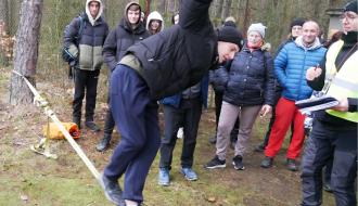 W ekwilibrystycznej pozie jeden z uczestników rajdu na taśmie slackline. Obok i z tyłu oglądający rywalizację piechurzy.