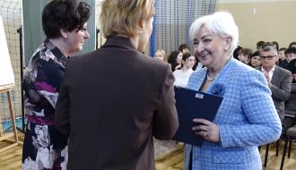 Uroczyste podsumowanie Forum w hali sportowej SP w Brzustowcu. Pani Wojewoda Łódzki w niebieskim żakiecie uśmiecha się, trzymając granatową teczkę, i patrzy na odwróconą tyłem Panią Dyrektor SP w Brzustowcu, ubraną w brązową marynarkę. Obok po lewej nauczycielka ubrana w kolorową bluzkę. W tle siedząca publiczność oraz elementy wyposażenia sali.