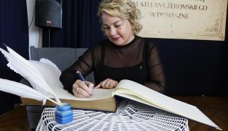Kobieta podpisująca księgę pamiątkową przy stole przykrytym ozdobnym, białym obrusem, na którym znajduje się kałamarz z białymi piórami. W tle baner informujący o nadaniu sztandaru szkole.