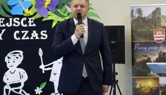  Senator Rafał Ambrozik w ciemnoniebieskim garniturze i krawacie przemawia do mikrofonu na tle dekoracji ściennej z postaciami dzieci i kwiatami.
