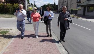 Grupa osób w strojach wyjściowych i codziennych spaceruje nowym chodnikiem z czerwonej kostki wzdłuż drogi. Na czele idą od lewej: I Wicewojewoda Łódzki, Wiceprzewodnicząca Rady Miejskiej w Drzewicy, Przewodnicząca Rady Miejskiej w Drzewicy, Burmistrz Drzewicy.