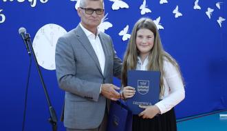 Burmistrz Drzewicy wręcza granatowy dyplom z herbem Drzewicy uśmiechniętej, długowłosej uczennicy.