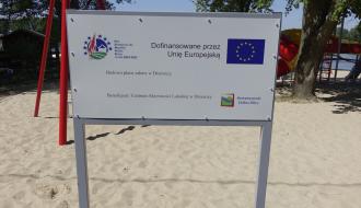 Biała tablica informacyjna na piasku, dokumentująca dofinansowanie budowy placu zabaw w Drzewicy przez Unię Europejską, z widocznymi unijnymi logotypami.