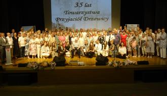 Liczna grupa uczestników i organizatorów stoi na scenie RCK podczas wspólnego, pamiątkowego zdjęcia. W tle na dużym ekranie wyświetla się napis „35 lat Towarzystwa Przyjaciół Drzewicy” oraz nagłówek „XIX Spotkanie po latach w Drzewicy”.