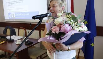 Sołtys Werówki, Zofia Danuta Kacprzak przemawia do mikrofonu na tle flag. W rękach trzyma duży, kolorowy bukiet kwiatów oraz wręczony wcześniej dokument.