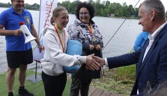  Oficjalne gratulacje nad wodą. Burmistrz Drzewicy w garniturze podaje rękę uśmiechniętej młodej kobiecie w jasnej bluzie, która trzyma niebieski pakunek. W tle stoi kobieta z medalem na szyi oraz mężczyzna w niebieskiej koszulce polo z megafonem w ręku. Za nimi widoczna jest flaga promocyjna Drzewicy.