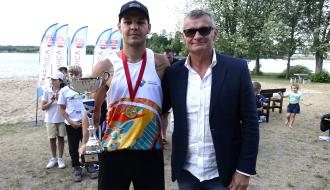 Pozowane zdjęcie młodego siatkarza z medalem i srebrnym pucharem dla najlepszego zawodnika turnieju oraz Burmistrza Drzewicy w marynarce i okularach. Z tyłu dzieci i flagi promocyjne Drzewicy. W tle plaża, jezioro i drzewa.