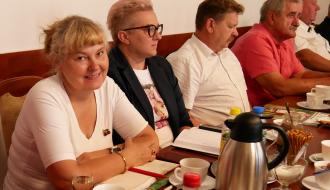 Uczestniczący w sesji sołtys siedzący przy stole obrad. Na pierwszym planie uśmiechnięta kobieta w białej bluzce z notatnikiem i długopisem, obok niej kobieta w krótkich jasnych włosach i okularach oraz mężczyźni w koszulach.