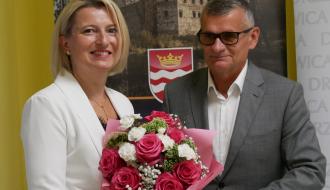 Dyrektorka żłobka w białym garniturze odbiera bukiet różowych i białych kwiatów od Burmistrza Drzewicy w szarym garniturze. W tle widoczna ścianka reklamowa Gminy Drzewica z herbem i zdjęciem zamku.