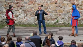 Burmistrz Drzewicy w czarnej kurtce i okularach przeciwsłonecznych przemawia do mikrofonu na scenie, stojąc między dwoma rekonstruktorami w strojach historycznych.