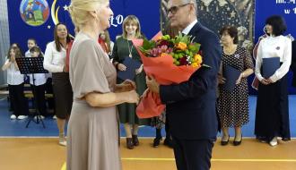 Burmistrz Drzewicy w garniturze wręcza duży, kolorowy bukiet kwiatów Pani Dyrektor SP w Radzicach Dużych, ubranej w beżowej sukience. W tle stoją pozostali uczestnicy uroczystości na tle niebieskiej dekoracji szkolnej. 