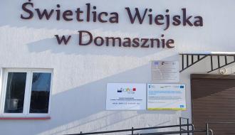 Frontowa ściana budynku z napisem „Świetlica Wiejska w Domasznie” oraz tablicami informacyjnymi