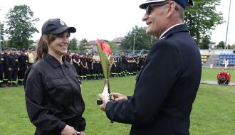 Burmistrz Drzewicy Janusz Reszelewski w galowym mundurze przekazuje złoty puchar za 1. miejsce uśmiechniętej druhnie OSP Domaszno w czarnym mundurze i czapce z daszkiem. W tle widać zgrupowane drużyny pożarnicze na tle trybun stadionu.