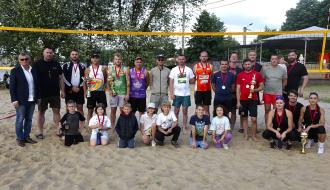 Oficjalne zdjęcie grupowe uczestników turnieju siatkówki plażowej z pucharami i medalami na tle siatki i jeziora.