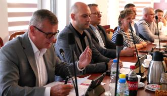 Burmistrz Drzewicy w okularach i siwym garniturze przemawia do mikrofonu, wykonując gest dłonią i przeglądając dokumenty na stole. Obok niego przy stole siedzą Wiceburmistrz i radni.