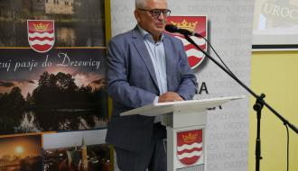 Wicewojewoda Łódzki w okularach i niebieskim garniturze przemawia przy mównicy ozdobionej herbem Gminy Drzewica. W tle znajduje się baner promocyjny z hasłem „Poczuj pasję do Drzewicy”.