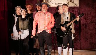 Grupa aktorów teatralnych w kostiumach z epoki występuje na scenie; w centrum mężczyzna w różowym surducie, obok niego mężczyzna gra na gitarze.