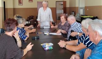  Kobieta stojąca na czele stołu i prowadząca spotkanie dla seniorów rozmawiających przy ciemnym stole w świetlicy w Domasznie. 