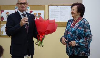 Burmistrz Drzewicy w ciemnym garniturze i okularach przemawia do mikrofonu i wręcza duży bukiet czerwonych kwiatów Pani Marii Staniszewskiej w wzorzystej bluzce. W tle na ścianie wiszą tablice korkowe z jadłospisem i pracami dzieci. 