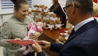 Uśmiechnięta matka z małym dzieckiem na rękach odbiera czerwoną teczkę. W tle stół z prezentami i pluszowymi misiami. 