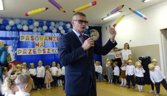 Burmistrz Drzewicy w garniturze i okularach przemawia przez mikrofon. Pod sufitem wiszą dekoracyjne, papierowe kredki.